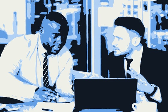 Two men in business dress talking across a laptop in a lobby.