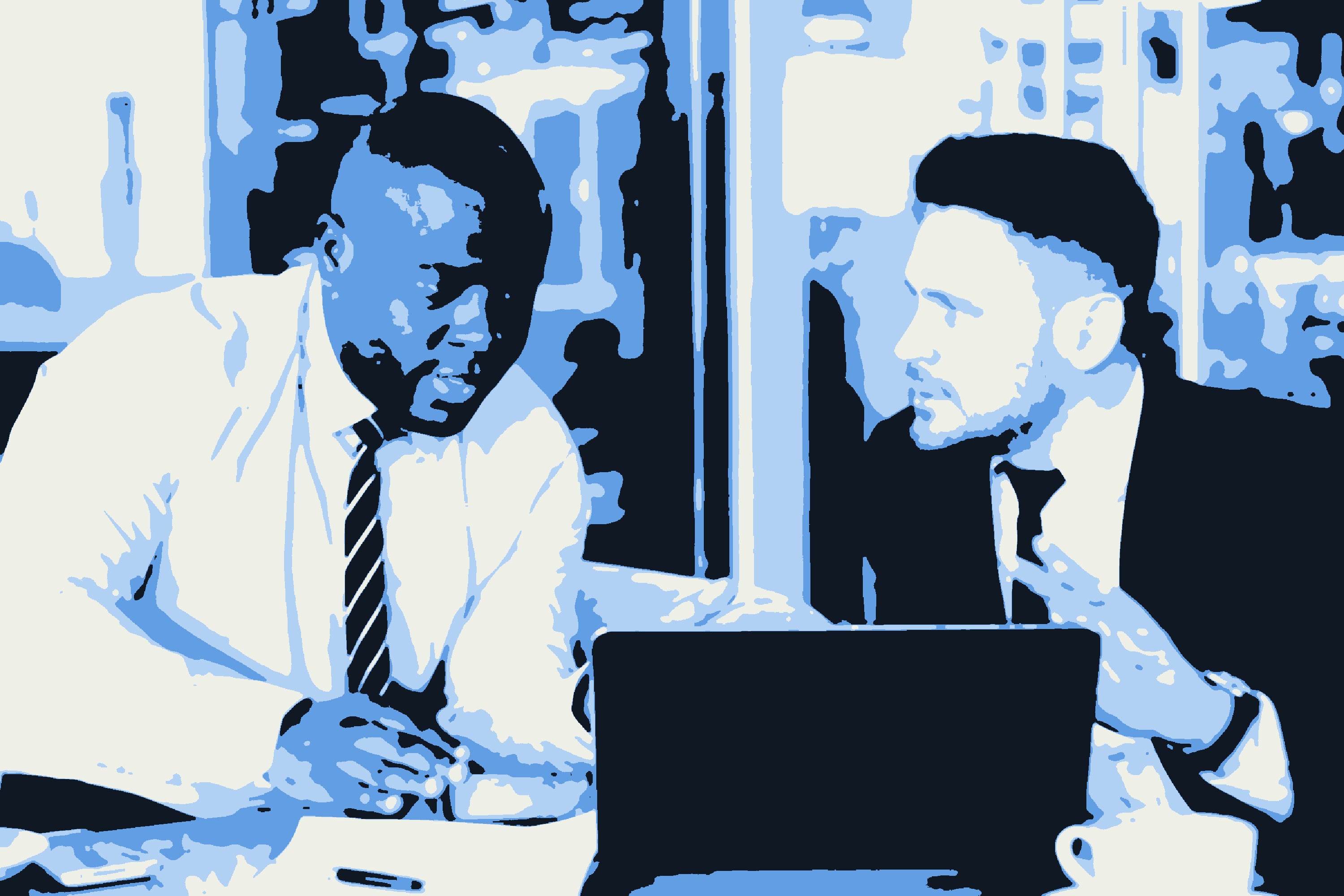 Two men in business dress talking across a laptop in a lobby.